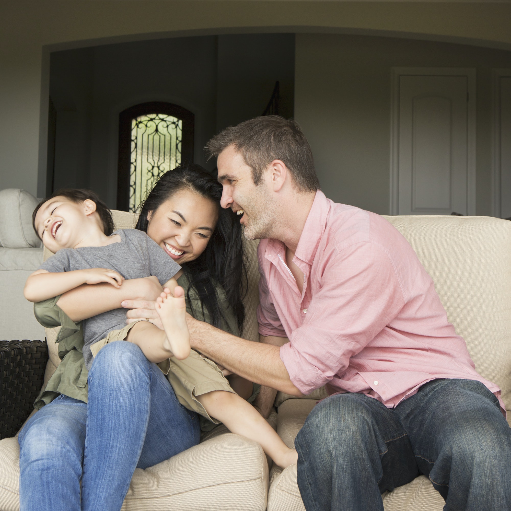 A family, a man and woman sitting side by side on a sofa playing with their young son.